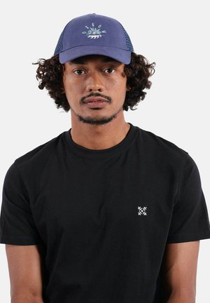 Homme aux cheveux bouclés portant une casquette bleue avec un motif de vague et un t-shirt noir uni avec un petit logo blanc sur la poitrine.