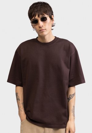 Νεαρό άτομο που φοράει σκούρα γυαλιά ηλίου, καφέ υπερμεγέθη μπλούζα, μπεζ παντελόνι, και εμφανίζει τατουάζ και στα δύο μπράτσα, πάνω σε απλό φόντο.