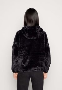 Persona con cabello largo y oscuro que lleva una chaqueta de capucha de felpa negra y pantalones negros, de pie de espaldas al espectador frente a un fondo sencillo.