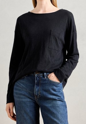 Vrouw met een zwart shirt met lange mouwen en borstzak, ingestopt in een blauwe jeans, met één hand in de zak van de jeans.