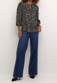 Bluse med blomsterprint i mørke farver og nøglehuludskæring, kombineret med blå jeans med vidde i benene og sorte spidse hæle.