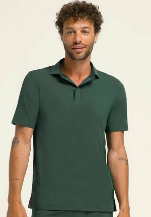 Uomo con capelli ricci e barba che indossa una polo verde scuro a maniche corte, in piedi davanti a uno sfondo chiaro e uniforme.