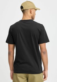 Černé bavlněné tričko s krátkým rukávem, standardní střih. Nenošeno s lehkými khaki šortkami, pohled zezadu, ukazující minimalistický design.