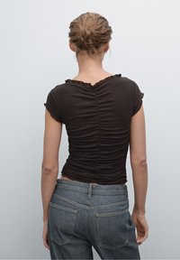 Brown, fitted top with short sleeves and ruched detailing on the back. Paired with mid-rise blue denim jeans featuring back pockets.