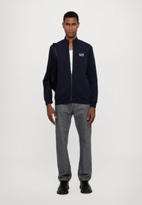 Felpa blu navy con zip e alto colletto, polsini a costine e logo EA7; abbinata a jeans grigi in denim e scarpe nere. Il modello porta una borsa nera.