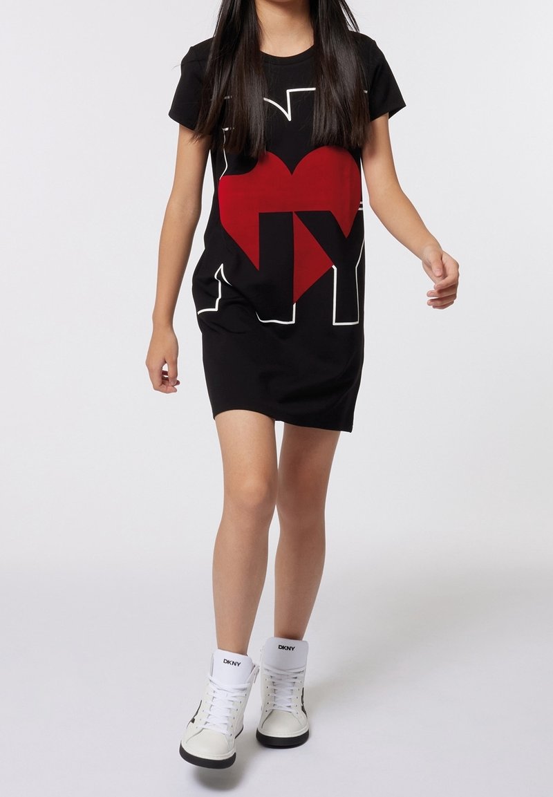 Fille marchant portant une robe noire avec un cœur rouge et l'inscription blanche « I NY », associée à des baskets montantes blanches.
