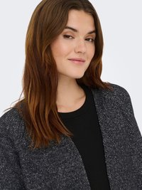 Young woman with long brown hair wearing a black top and charcoal gray knit cardigan, smiling slightly, against a plain light background.