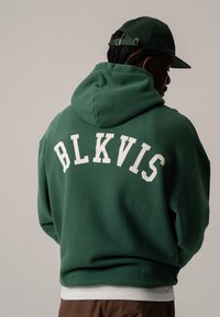 Hombre con sudadera verde y gorra a juego, de espaldas a la cámara, texto "BLKVIS" en blanco en la sudadera, fondo neutro.