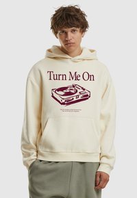 Cremefarbener Kapuzenpullover mit einem Grafikmotiv eines Plattenspielers und dem Text "Turn Me On" in Burgunderrot, mit einer frontalen Tasche und gerippten Bündchen.