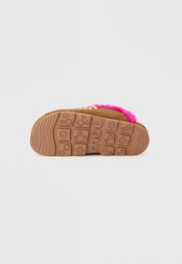 Semelle brune de chaussure avec des motifs, montrant le nom de la marque brodé, et un dessus en tissu peluche rose visible sur le bord supérieur.