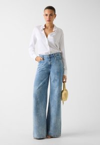 Heleen sinised, kõrge vöökohaga teksapüksid laia säärega ja tuhmiks kulunud tekstuuriga; kombineeritud valge nööpidega särgiga ja käes hoides väikest kuldset kotti.