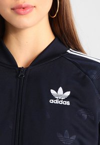 Svart zip-up jacka med hög krage, prydd med vitt Adidas-logotyp och tre vita ränder på axlarna, tillverkad av slätt, strukturerat tyg.