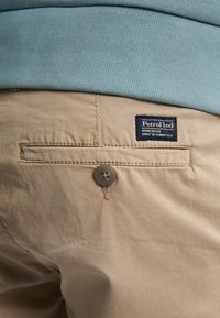 Beige katoenen shorts met een achterzak met een bruine knoop en een blauwe label waarop staat "Petrol Ind, Denim Driven, sinds '69 Florida U.S.A."