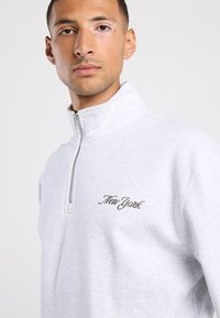 Felpa a zip di colore grigio chiaro con colletto alto; presenta la scritta ricamata "New York" in un font corsivo sulla parte sinistra del petto.