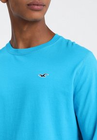 Light blue cotton crew neck t-shirt featuring a small black bird logo on the left chest and visible stitching details.