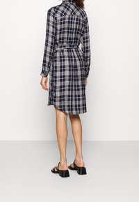 Donna che indossa un abito camicia a quadri blu navy e bianco con cintura, maniche lunghe e sandali con tacco neri, in piedi di spalle su uno sfondo neutro.