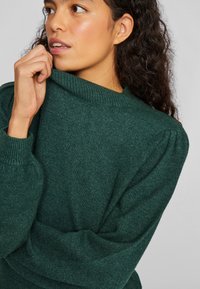 Groene gebreide trui met een hoge, geribbelde kraag, gestructureerde stof en geplooide pofmouwen. Het ontwerp heeft een losse pasvorm en zachte materialen.