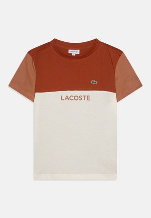 Camiseta de manga corta con un bloque de color marrón en la parte superior, sección inferior color crema y texto "LACOSTE". Hecha de material de algodón suave.