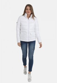 Veste blanche duveteuse avec un col haut, fermeture éclair à l'avant, deux poches latérales et une texture lisse et brillante. Portée avec un jean bleu et des baskets blanches.