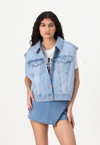 Gilet in denim azzurro chiaro con colletto, chiusura a bottoni e due tasche sul petto. Indossato sopra una t-shirt bianca con grafica e una gonna in denim blu.