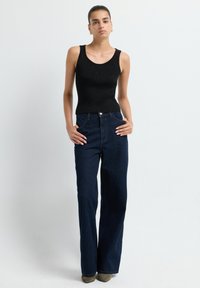 Top sin mangas negro con relieve combinado con vaqueros acampanados de cintura alta en azul oscuro. La modelo está de pie con las manos en las caderas, mostrando el atuendo.