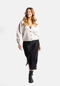 Kremowy, oversize'owy kardigan z drewnianymi guzikami, w zestawieniu z czarną satynową spódnicą na ramiączkach z bocznymi rozcięciami oraz czarnymi, sznurowanymi butami typu combat.