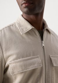 Beige corduroy jas met een ritssluiting aan de voorkant, een platte kraag en twee borstzakken. Heeft een verticale ribbelstructuur en zilveren ritshardware.