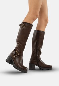 Botas de cuero marrón hasta la rodilla con un tacón grueso, detalles decorativos de hebillas y cremallera lateral. Suela texturizada para mayor agarre.