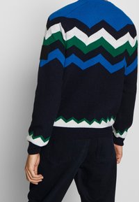 Knitted sweater featuring a zigzag pattern in blue, green, white, and navy. Ribbed cuffs and hem; includes a black patch on the shoulder.