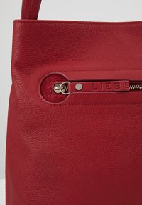 Sac en cuir rouge en gros plan montrant une surface texturée et une languette de fermeture éclair embossée du mot "LIEBE" en détail métallique.