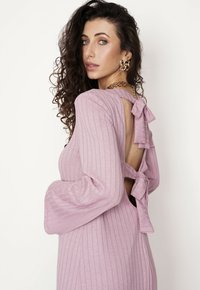 Zaszewek sukienka z długim rękawem w jasnym różu z wycięciem z tyłu, które jest zabezpieczone dwoma kokardkami. Tkanina ma miękką teksturę i dobrze dopasowuje się do sylwetki.