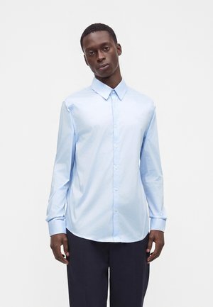 Jeune homme portant une chemise bleu clair boutonnée et un pantalon foncé, debout sur un fond blanc uni.
