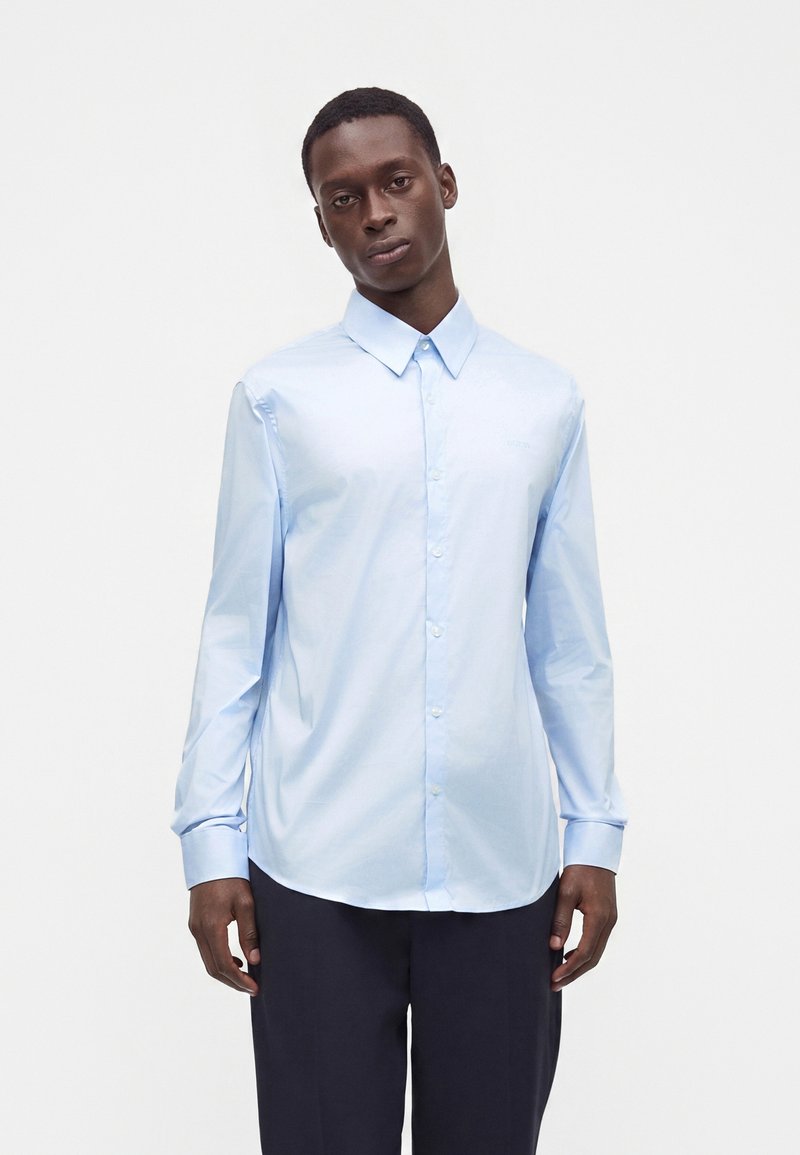 Jeune homme portant une chemise bleu clair boutonnée et un pantalon foncé, debout sur un fond blanc uni.