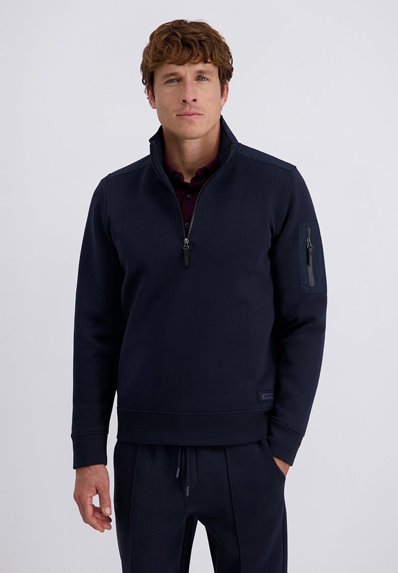 Mann in navyblauer Quarter-Zip-Pullover mit einer Reißverschlusstasche am Ärmel und passender navyblauer Hose vor einem schlichten Hintergrund.