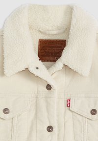 Giacca in corduroy crema con colletto bianco peloso, etichetta in pelle marrone, bottoni metallici e due tasche sul petto.