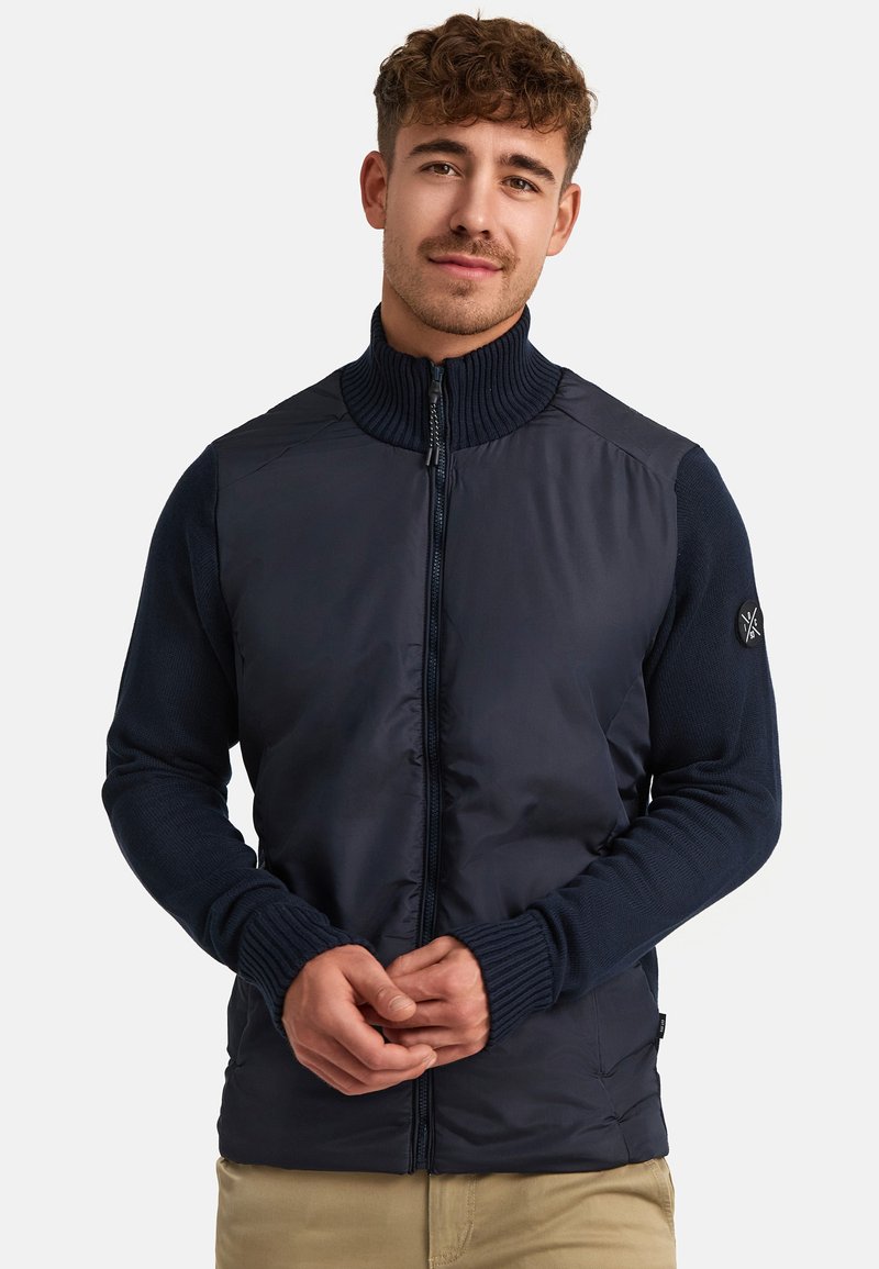 Navy jacket with a quilted front and knitted sleeves. Features a high collar, front zipper, and a logo patch on the left sleeve.