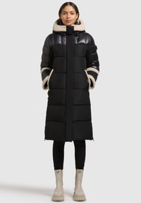 Sort puffjakke med hætte og beige pelskant. Jakken har et geometrisk quiltet design og forlommer. Båret over sorte leggings og cremefarvede støvler.