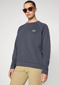 Mulher vestindo um moletom cinza escuro com um pequeno logotipo colorido no peito, calças bejes e óculos de sol tartaruga, em pé contra um fundo plain.