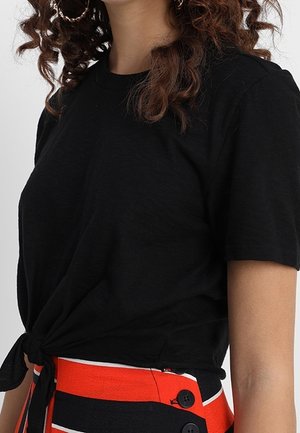 Vrouw met krullend haar draagt een zwart T-shirt met korte mouwen, geknoopt in de taille, en een hoge taille rok met rode, zwarte en witte horizontale strepen met knopen.