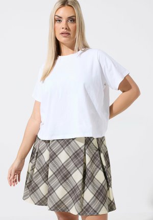 Blondynka w białej, gładkiej koszuli z krótkim rękawem i beżowo-szarej plisowanej spódnicy w kratę na białym tle.