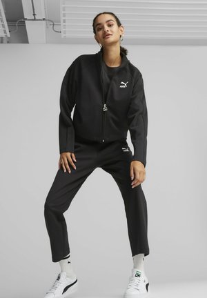 Tuta sportiva nera con giacca con zip e pantaloni affusolati. Presenta il logo Puma sul petto e sulla coscia. Abbinata a sneakers e calzini bianchi.