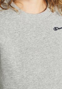 Sudadera gris con cuello redondo. Confeccionada en suave material de algodón, cuenta con un pequeño logo de Champion en color azul oscuro en el pecho.