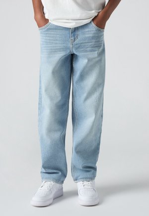 Personne portant un jean bleu clair coupe large, des baskets blanches et une chemise blanche, les mains dans les poches, sur un fond uni.