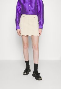 Jupe beige à bords festonnés avec une texture ondulée et des boutons dorés, associée à une chemise violette en satin brillant et des chaussettes noires avec des chaussures noires.