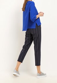 Blue blouse with three-quarter sleeves, paired with navy pinstripe trousers and silver sneakers, showing a modern, relaxed fit.