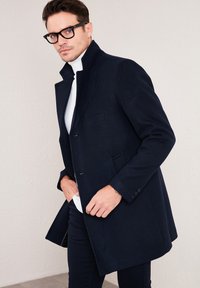 Abrigo de lana azul marino con cuello alto, diseño de un solo botón, dos bolsillos frontales y textura suave, combinado con un suéter blanco de cuello alto.