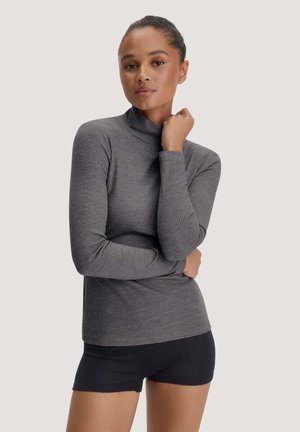 Femme aux cheveux foncés en chignon, portant un pull gris côtelé à col roulé manches longues et un short noir, debout les bras croisés.