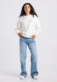 White sweatshirt with a small yellow lemon embroidery, paired with light blue straight-leg jeans and white sneakers. Flat texture, casual fit.