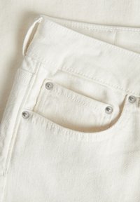 O tecido de denim branco possui um bolso com rebites de metal prateado. A textura apresenta um padrão de sarja visível, com detalhes de costura evidentes.