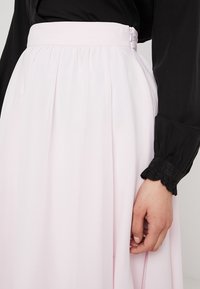 Falda rosa claro, fluida, con pliegues y cremallera lateral. Combinada con una blusa negra de manga larga con puños fruncidos.